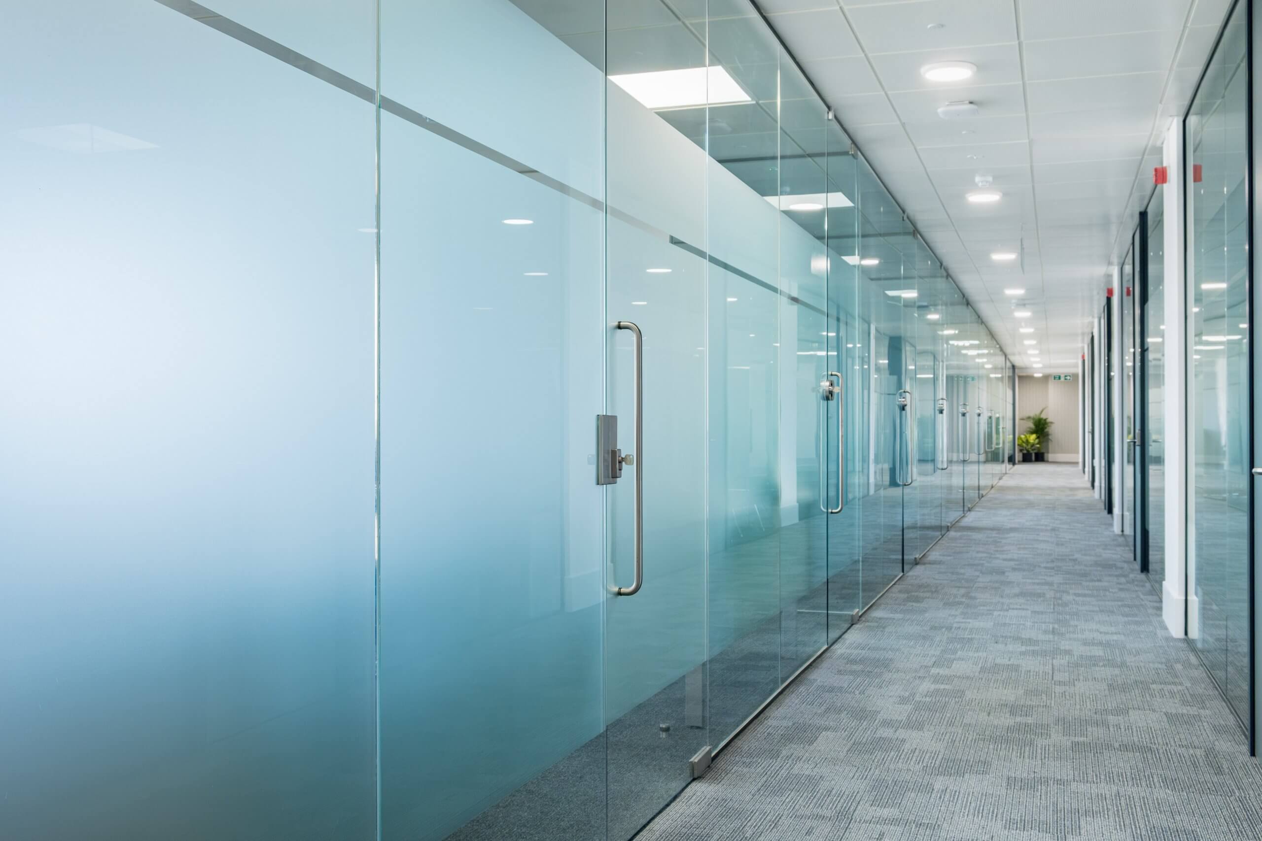 Office corridor with rows of offices on either side, separated by glass partitioning.